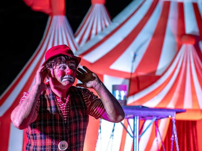 A clown in a red check waistcoat in front of the red and white circus tent
